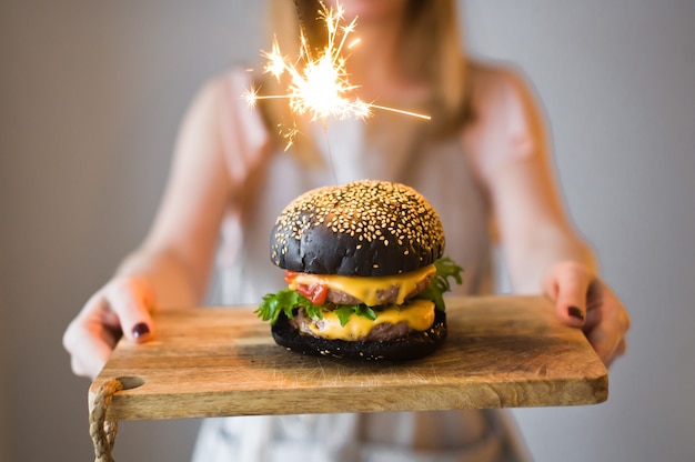 The chief is holding a wooden cutting Board with a black Burger.