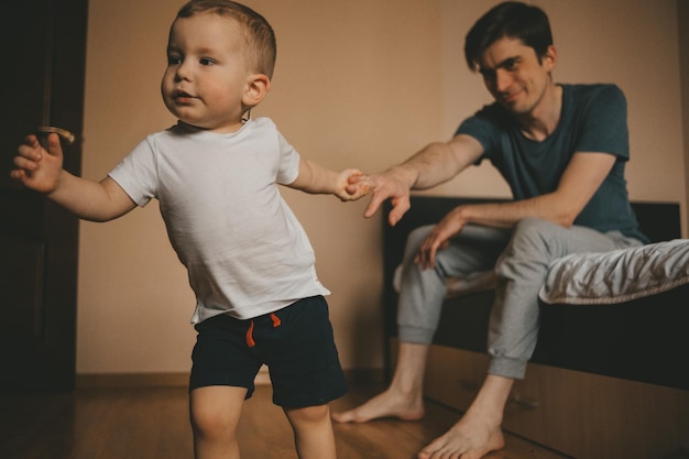 Child of 2-3 years old holds his father\'s hand and leads him\
forward. dad is sitting on the bed