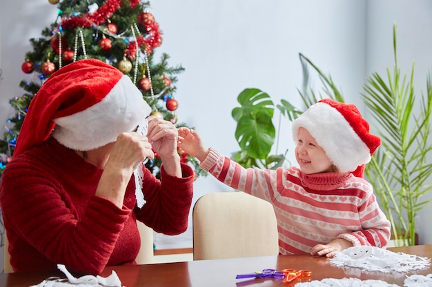 サンタクロースの帽子をかぶった子供と大人は、クリスマスに家を飾るためにクリスマスの装身具を作ります。おばあちゃんとの幸せな休日とクリスマス。