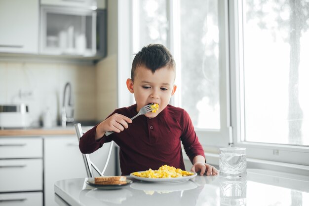 A child in the afternoon on a white light kitchen in a burgundy sweater eats an omelet