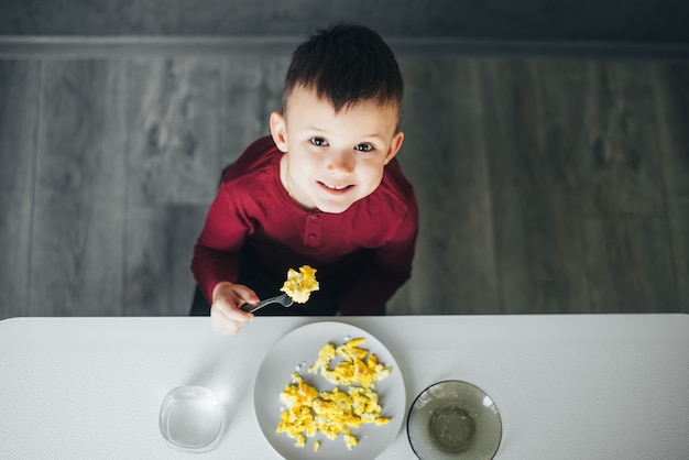 A child in the afternoon on a white light kitchen in a burgundy sweater eats an omelet