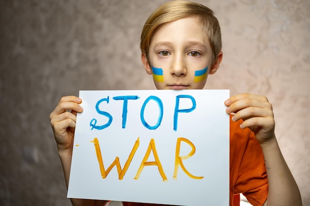 A child against war a boy with a painted flag of Ukraine on his cheeks holding a poster with the inscription stop the war