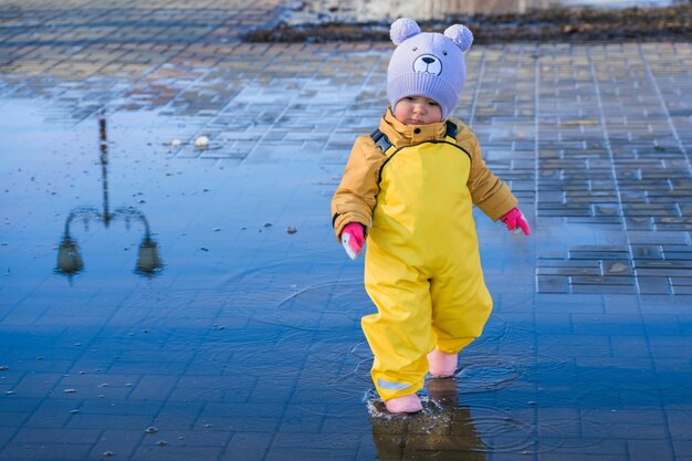 A child aged 23 years old dressed in a yellow rubber jumpsuit walks through puddles in the park