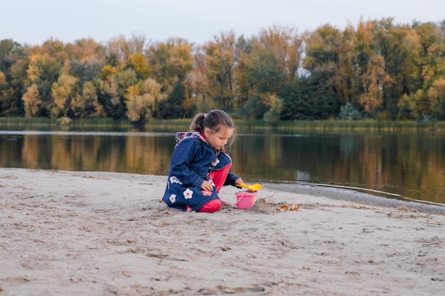 Child in autumn clothes is playing on beach little girl makes cakes out of sand on river bank on bac...