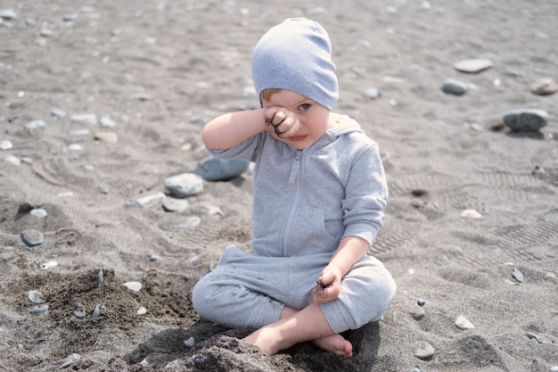 Детский белокурый мальчик играет с камнями и песком на пляже в солнечный день весной или осенью