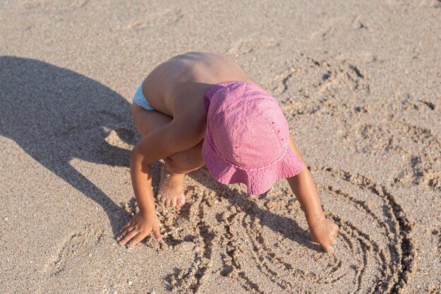 Child blond girl draws on the sand on the beach. Top view, flat lay.