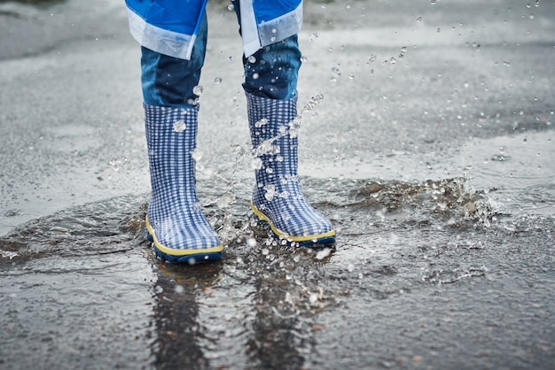 Bambino in stivali di gomma blu che salta sopra la pozzanghera sotto la pioggia