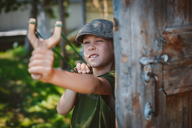 Child boy in a cap takes aim with a slingshot to shoot at a target. Play around as a child in the village on vacation.