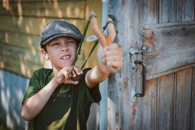 Ребенок мальчик в кепке прицеливается с рогаткой, чтобы стрелять в цель. Играем в детстве в деревне на отдыхе.
