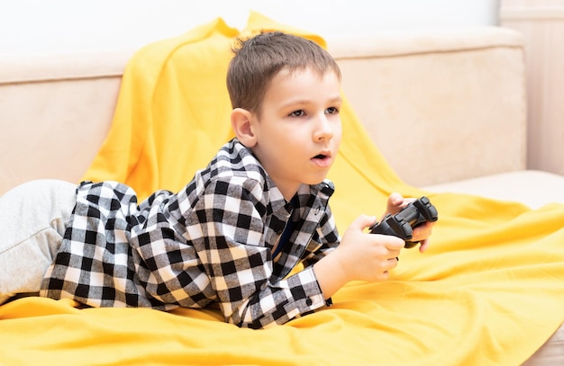 The child boy in checked shirt lying on the couch with black joystick in his hands playing the video game Playing video games at home concept