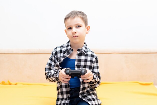 The child boy in checked shirt sitting on the couch with black joystick in his hands playing the video game Playing video games at home concept