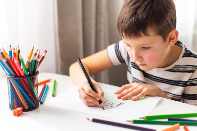 아이 소년 은 테이블 에 앉아 있는 동안 색 의 연필 으로  종이 에 그림 을 그린다