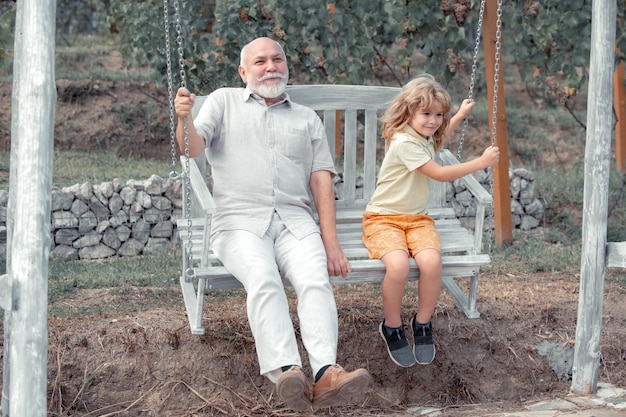 子供の男の子と祖父は夏の庭でスイングしますおじいちゃんと孫は公園でブランコに座っています
