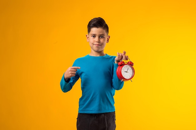 Photo child boy holding alarm clock and pointing with finger at it over yellow background time management concept