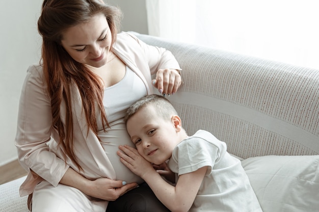 Ребенок мальчик обнимает живот беременной ее матери на диване у себя дома.