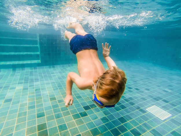 A child boy is swimming underwater in a pool, smiling and holding breath, with swimming glasses