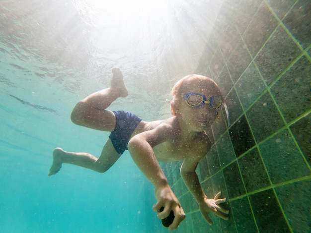 子供の男の子がプールで水中を泳いで、笑顔で息を止めて、水泳用メガネをかけています