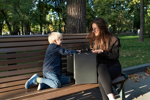 Child boy and nanny are sitting on bench in park and considering shopping bags