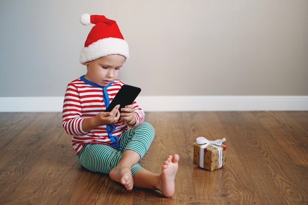 그의 손에 전화로 새해 복을 입은 아이 소년은 선물 옆에 바닥에 앉아있다.
