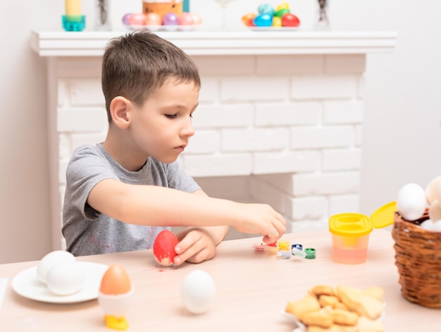 Child boy painting easter eggs against defocused fireplace with Easter decorations on it Selective focus Happy Easter concept Process of crafting at home