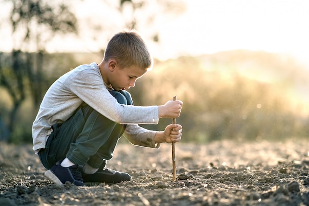 Child boy playing with wooden stick digging in black dirt ground outdoors