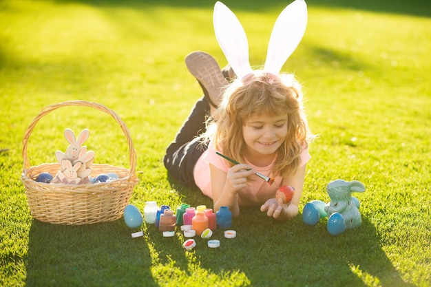 Child boy in rabbit costume with bunny ears painting easter eggs on grass in spring park