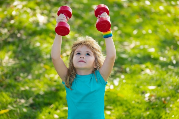 Child boy raising a dumbbell Cute child training with dumbbells Children fitness Kid boy exercising with dumbbells outdoor Healthy childhood lifestyle Sport strong boy little bodybuilder