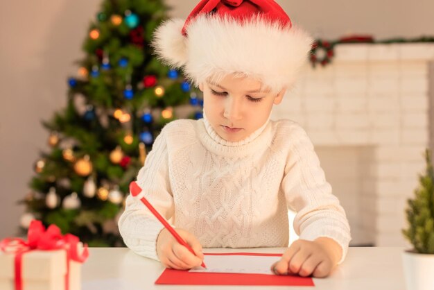 赤いサンタの帽子をかぶった子供がサンタクロースのクリスマスや新年の居心地の良い休日に手紙を書いています