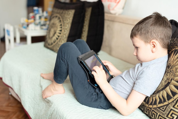 Child boy sitting on the coach with tablet playing games or studying something on it Social and technology concept