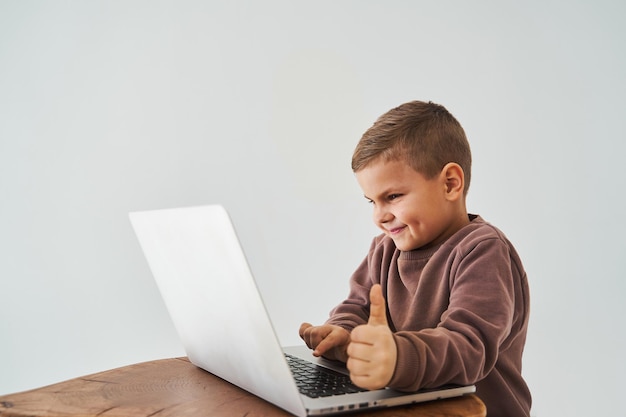 어린 소년은 노트북으로 온라인 공부를 하고 엄지손가락을 치켜세우고 웃는 모습을 보여줍니다. 온라인 교육 및 전자 학습 노트북으로 온라인 과정을 수강하는 어린이