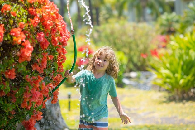 Child boy watering the plants from hose spray with water hose in the garden at the backyard of the house on a summer evening