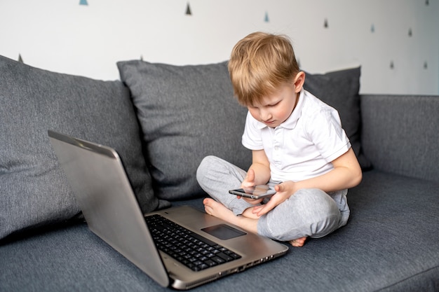 Child boy in a white t-shirt sits on a gray sofa and plays on a laptop