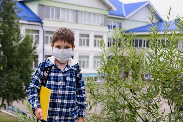 Child boy with bag, mask and book stays near elementary school