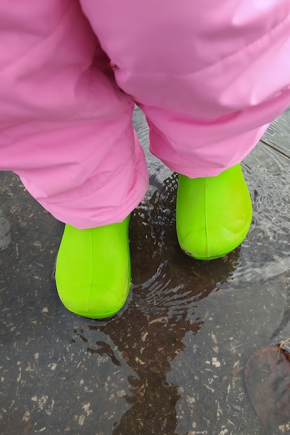 A child in bright green polyurethane boots and pink pants stands in an autumn puddle