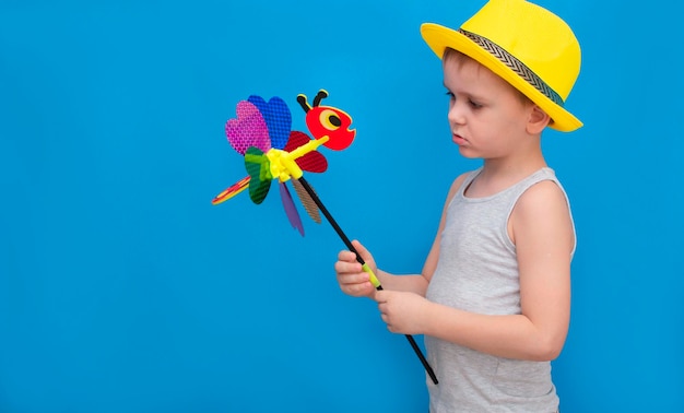 Ребенок в ярко-желтой летней шляпе держит блестящий игрушечный ветерок, стоящий на синем фоне. Мальчик наслаждается летними каникулами. Концепция счастливого детства.