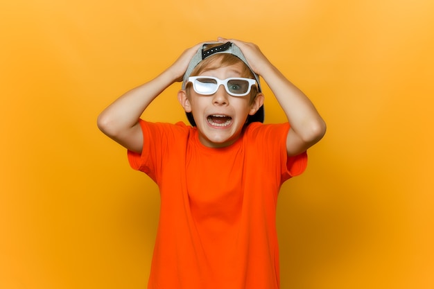 Photo a child in a cap and glasses for watching movies grabbed his head with both hands and shows emotions of fear