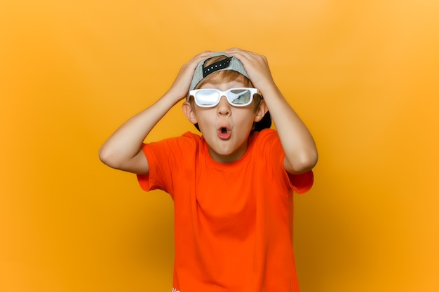 A child in a cap and glasses for watching movies grabbed his head with both hands and twisted his lips into a tube and shows emotions of surprise