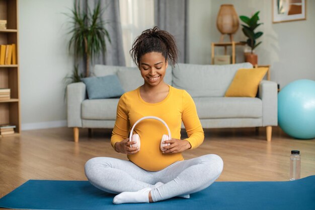 보육 매트에 앉아 큰 배꼽에 이어폰을 적용 쾌활한 젊은 임신 아프리카계 미국인 여자