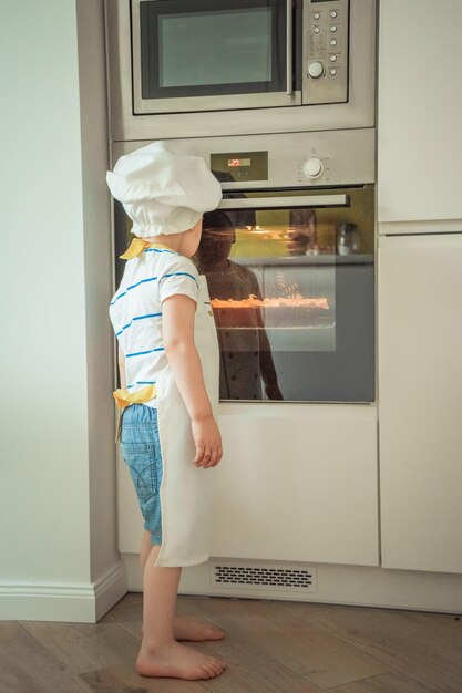 A child in a chef hat stands in front of an oven.