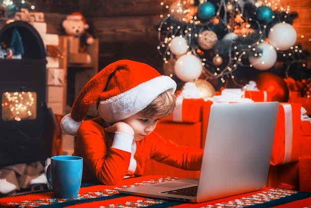 Ребенок у рождественской елки и у камина серфинг в Интернете Ребенок покупает рождественские подарки онлайн
