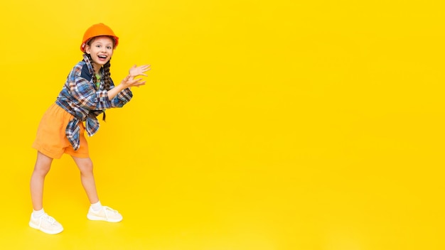 A child in a construction helmet points to your advertisement happy little teen girl in full height a place to advertise yellow isolated background copy space banner