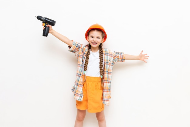 A child in a construction helmet and with a drill in his hands on a white isolated background A little girl with pigtails is preparing for repair by spreading her arms in different directions