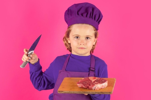 Child cook hold cutting board with meat beef steak and knife Excited chef cook Child wearing cooker uniform and chef hat preparing food studio portrait