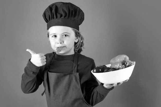 Child cook hold plate with vegetable Portrait of little child in uniform of cook Chef boy isolated on studio background Cute child to be a chef Child dressed as a chef hat