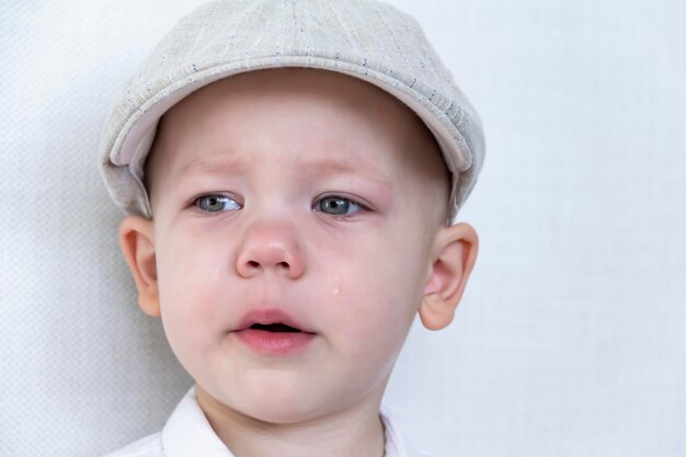 A child cries with eyes full of tears in a white shirt and cap near a white wall A crying little boy