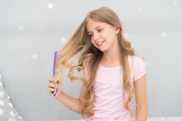 子供の巻き毛の髪型は、ヘアブラシまたは櫛を保持します。髪をとかす前にオイルを塗ってください。健康な髪。コンディショナーまたはマスク有機オイルコームヘア。ビューティーサロンのヒント。女の子の長い巻き毛の灰色のインテリアの背景。
