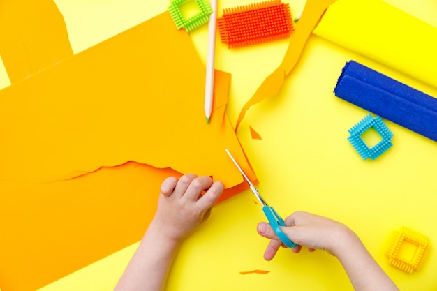 Child cutting colored orange paper with scissors  on a table for some craftwork