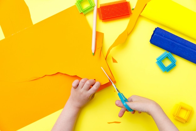 Child cutting colored orange paper with scissors on a table for some craftwork