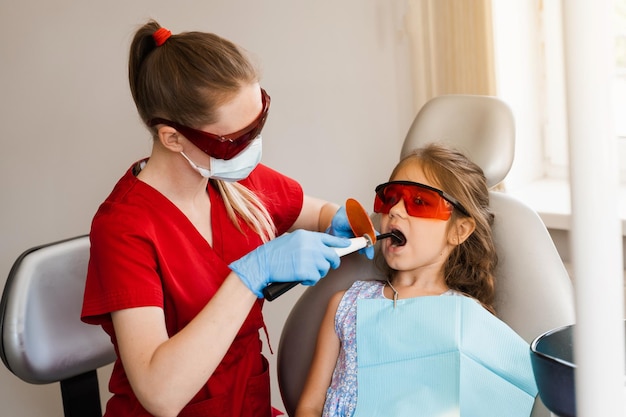 Child dentistry Uv illumination of photopolymer tooth filling procedure Child dentist in red protective glasses treats and removes caries in a patient