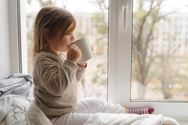 子供は大きな白いマグカップから窓の近くでココアまたは牛乳を飲みます